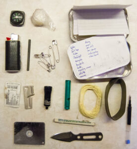 The contents of my kit. Top left is the compass and tinder (vaseline soaked cotton ball wrapped in plastic). Below that is mini BIC lighter wrapped in duct tape, a thin firesteel, and safety pins. Second row from the bottom, from left to right, has: a single Ondansetron anti-nausea pill, a P-38 can opener, a LED flashlight, whistle, kevlar string, ranger band, and pen. Bottom has signal mirror, roll of cash, and knife. Top right has the tin, with two pieces of waterproof blank paper and a packing list that fits in the lid.